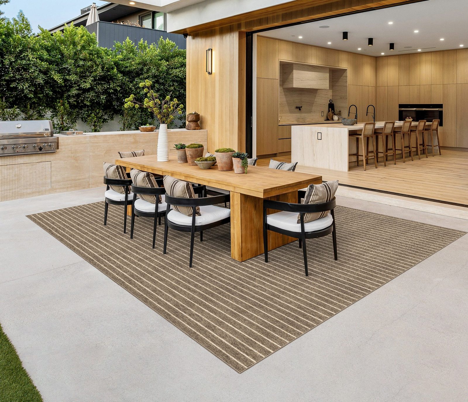 Striped taupe-and-cream outdoor flatweave rug under a teak dining table and black chairs on a concrete patio beside an outdoor kitchen
